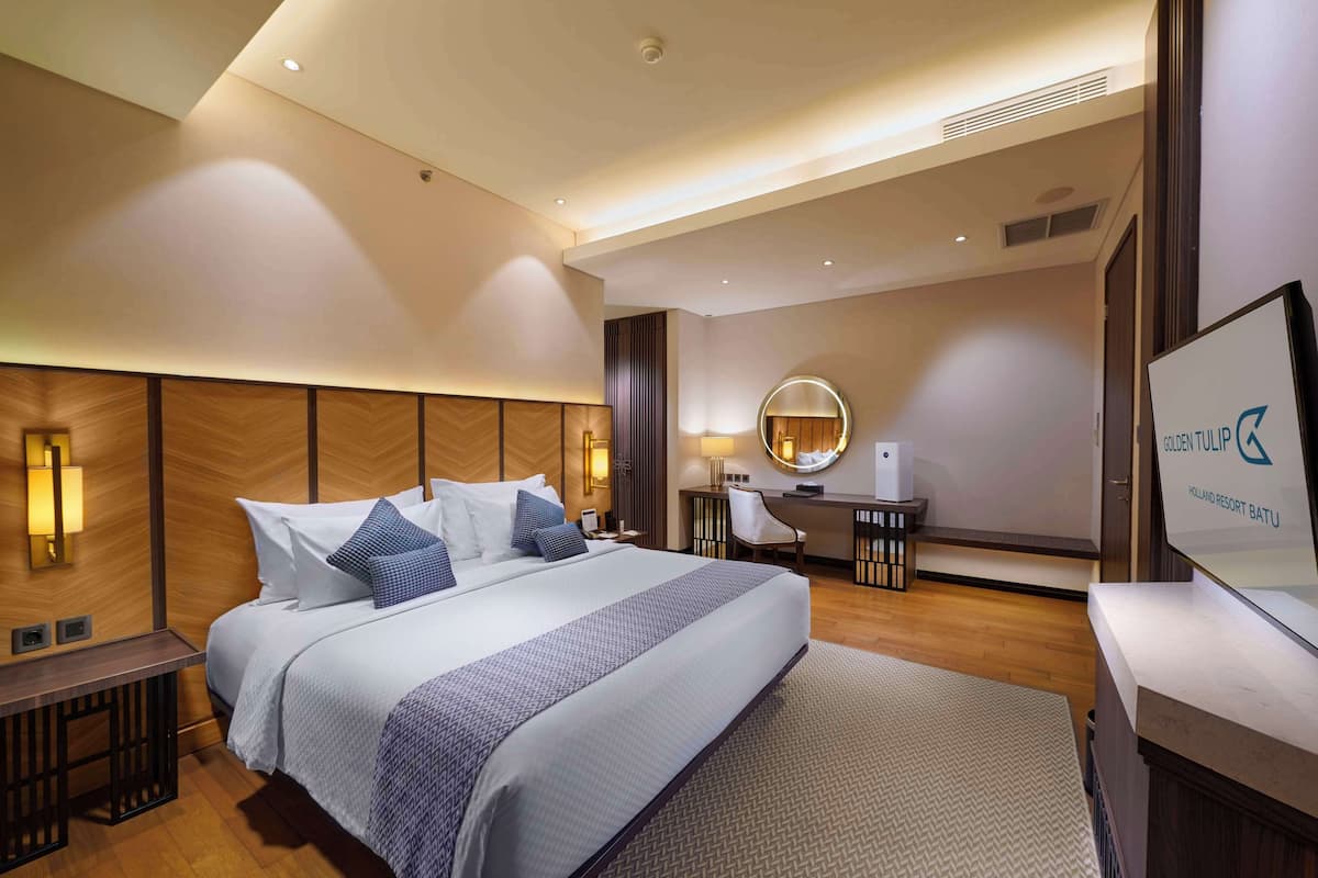 A modern hotel room with a large bed, a desk, a chair, a round mirror, and a TV.