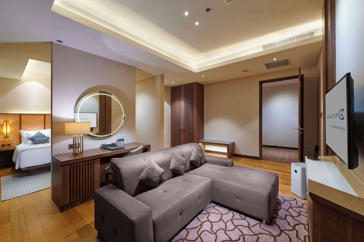 A modern hotel room with a large L-shaped sofa, a desk with a lamp, a round mirror, and a television.