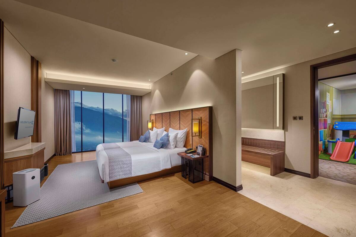 A modern hotel room with a large bed, a TV, a desk, and a view of the mountains.