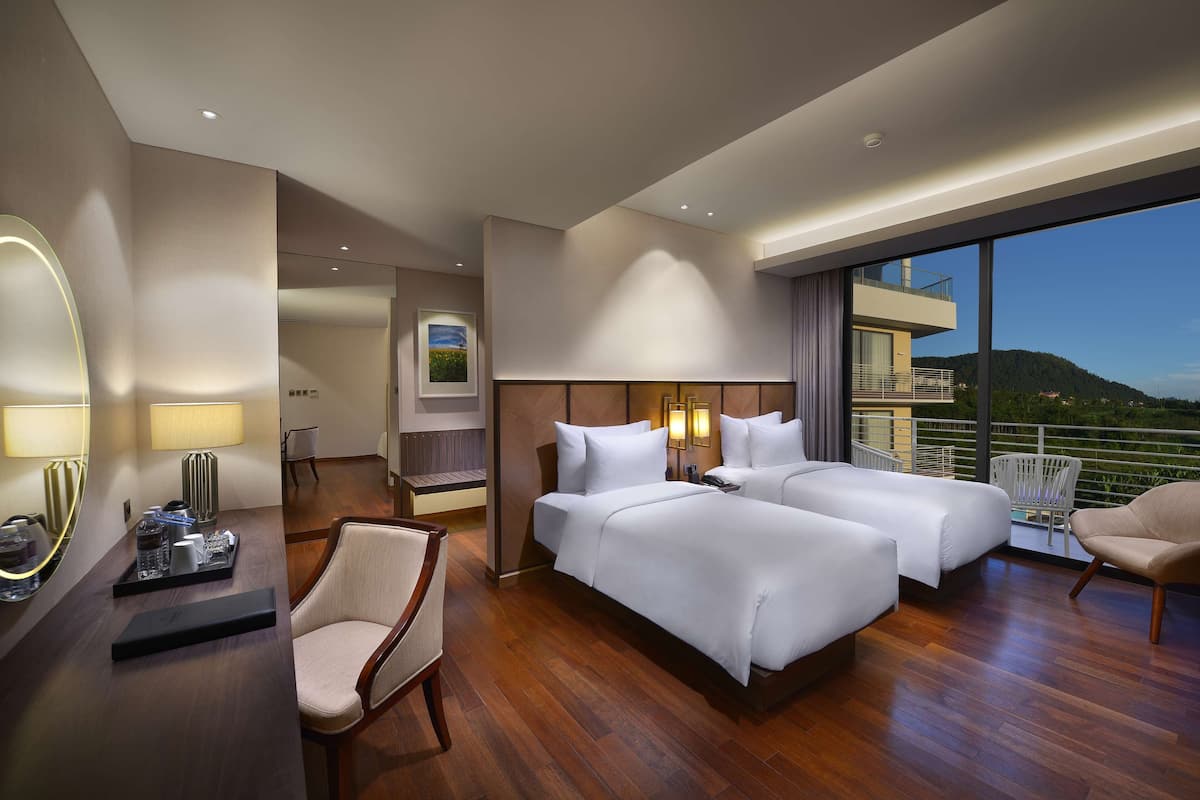 A hotel room with two beds, a desk with a lamp, a chair, and a balcony with a view of the mountains.