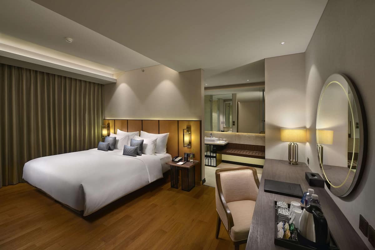 A hotel room with a large bed, bedside table with a phone and lamp, a chair, a desk with a mirror, and a bathroom with a sink and mirror.