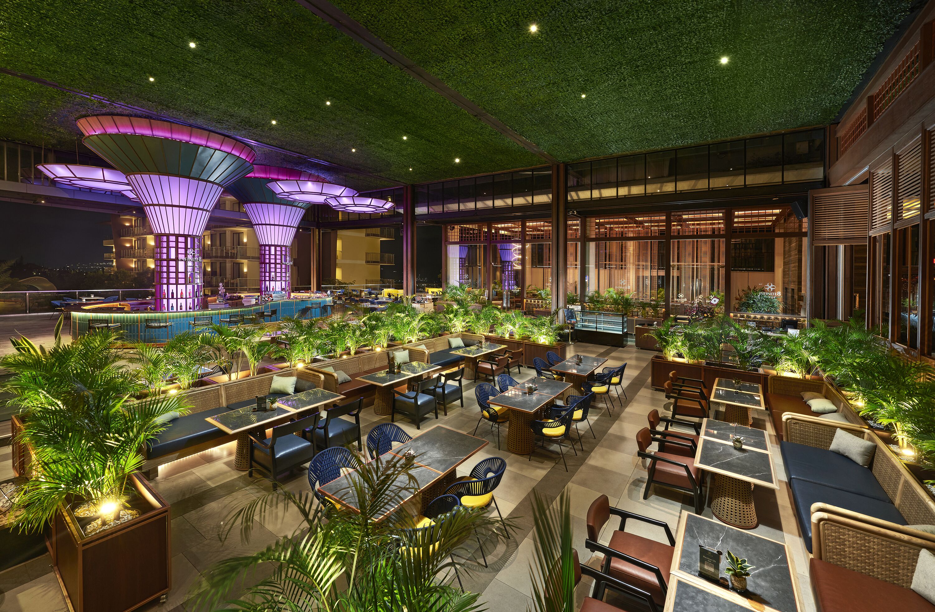 An indoor restaurant with multiple seating areas, including tables and booths, surrounded by potted plants.
