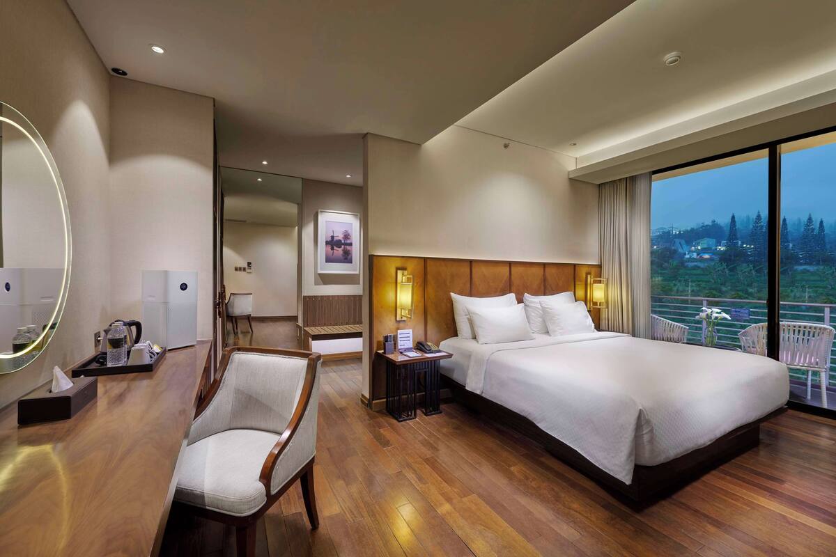 A hotel room with a large bed, a chair, a bedside table, a mirror, and a view of the outdoors.