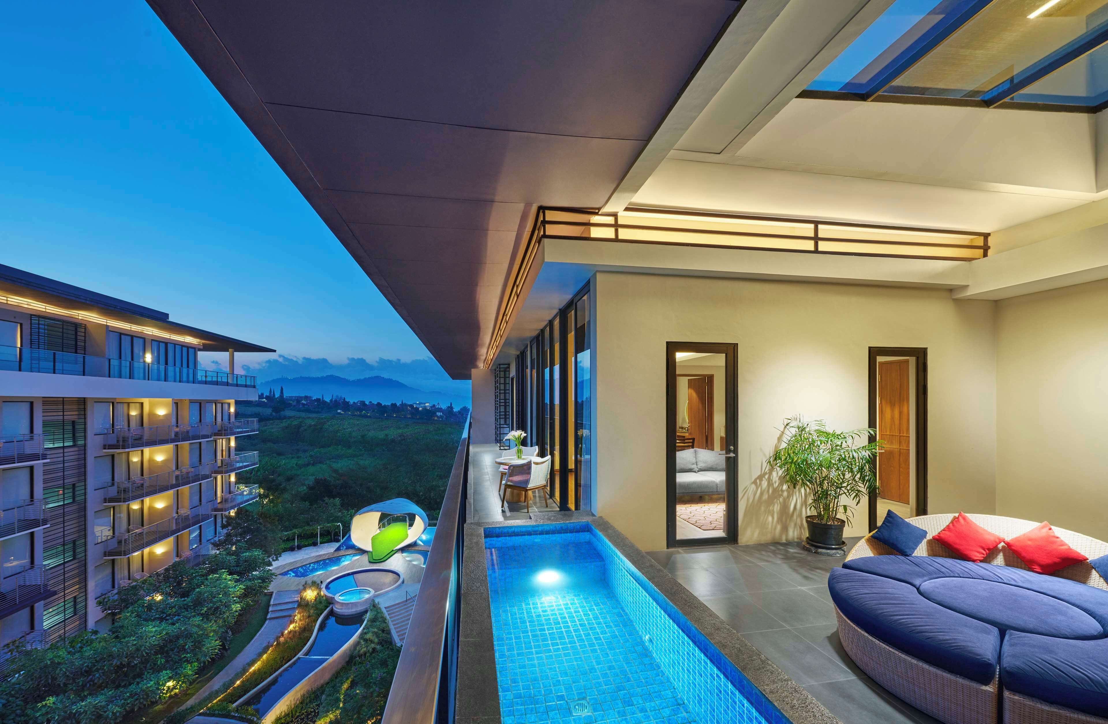 Un balcón de hotel moderno con piscina, mobiliario exterior y vistas del paisaje circundante.