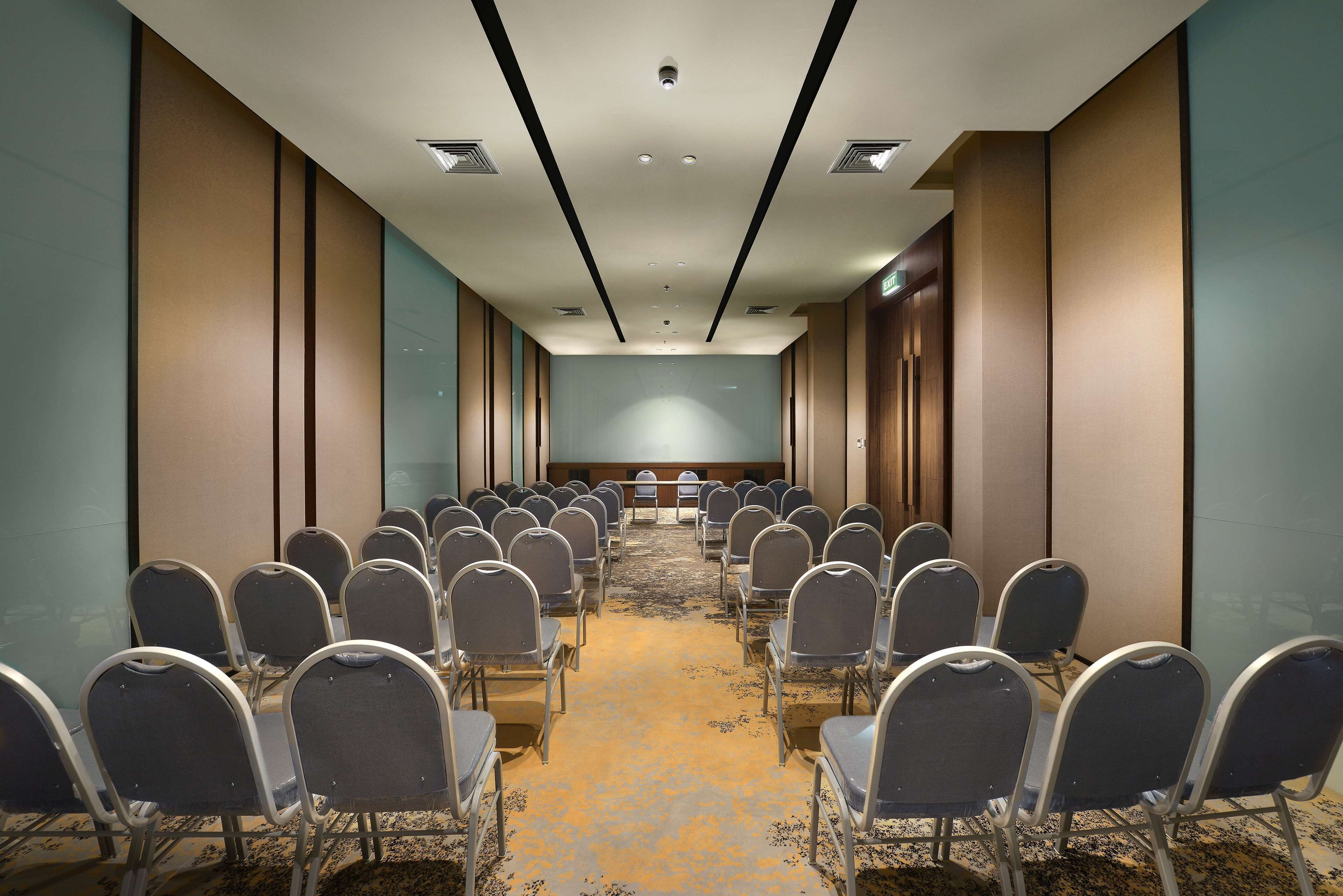 A conference room with rows of chairs facing a long table, a carpeted floor, and a high ceiling with recessed lighting.