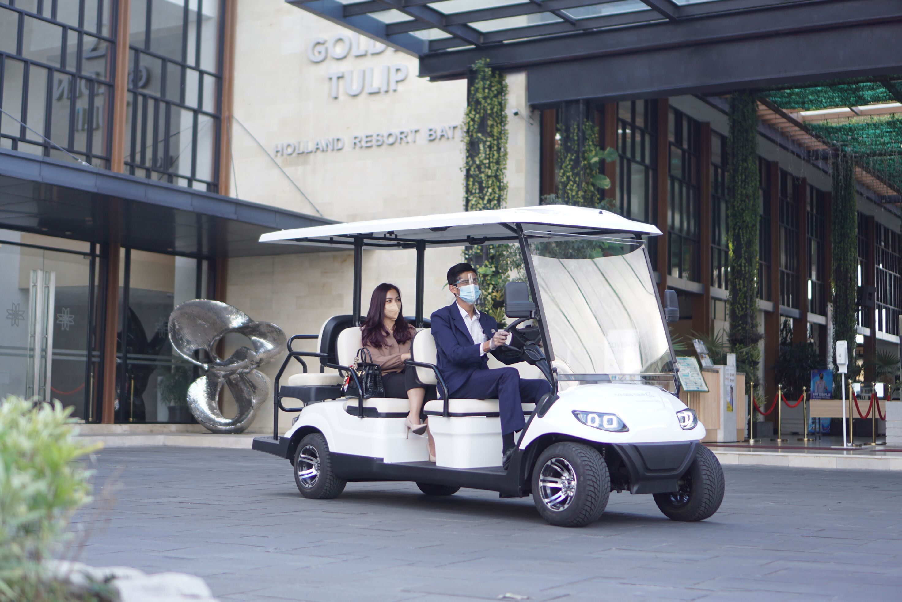 A golf cart with two passengers in front of a building with a sign reading "GOLD TULIP HOLLYWOOD RESORT BATH".