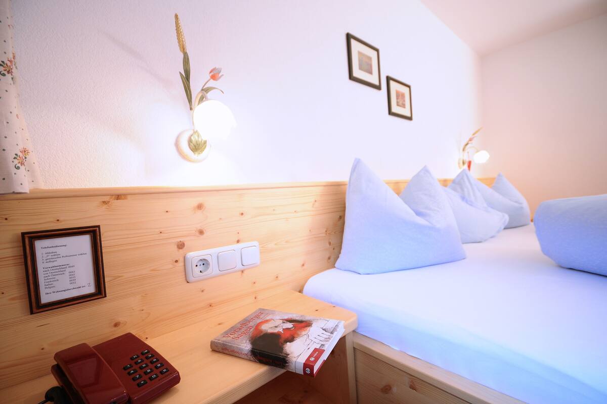 Habitación de hotel con cabecera de madera, mesita de noche con teléfono y revista, cama con ropa blanca y almohadas, y lámpara fijada a la pared.