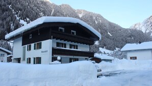 A multi-story building with a snow-covered roof, surrounded by snow-covered mountains. - Ferienwohnung Ahnenhöfl (Gaschurn)