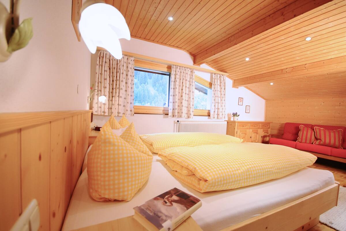 Una habitación acogedora con techo de madera, una cama con ropa de cama a cuadros amarillos, un sofá rojo y una ventana con cortinas.