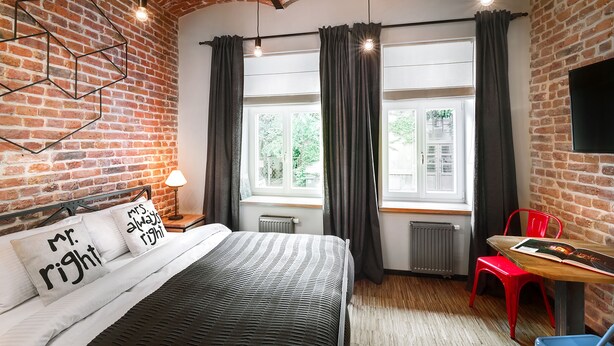 A room with a brick wall, a bed with a striped bedspread, two bedside lamps, a window with curtains, a radiator, a red chair, and a table with a book. - Well Well Aparthotel (Kraków)