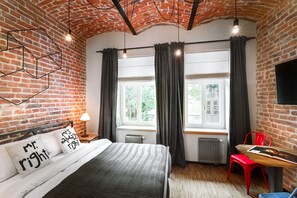 Een kamer met een bakstenen muur, een bed met een gestreept dekbedovertrek, twee bedlampen, een raam met gordijnen, een radiator, een rode stoel en een tafel met een boek.