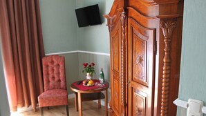 A room with a wooden cabinet, a red upholstered chair, a small table with fruit and a vase, and a flat-screen TV mounted on the wall.