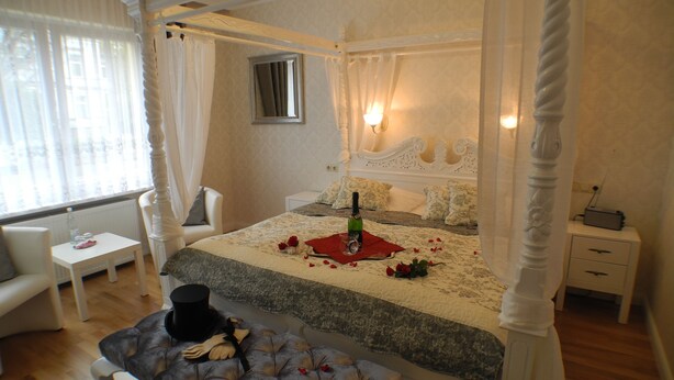 A four-poster canopy bed with a bottle and glasses on a red cloth, a tufted bench, a bedside table, and a nightstand with a radio.