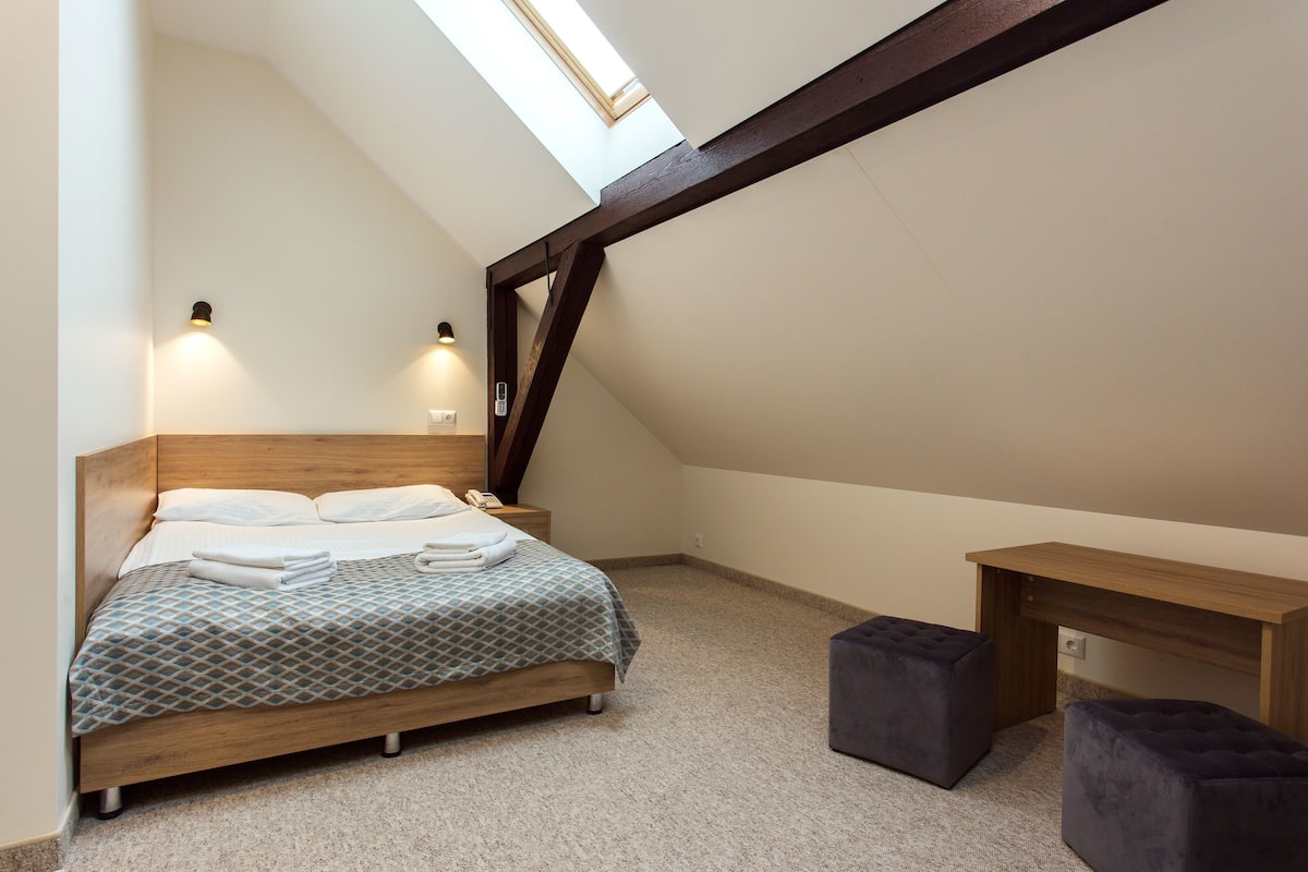 A bedroom with a sloped ceiling, a bed with a patterned bedspread, two bedside lamps, and a small desk with a chair.