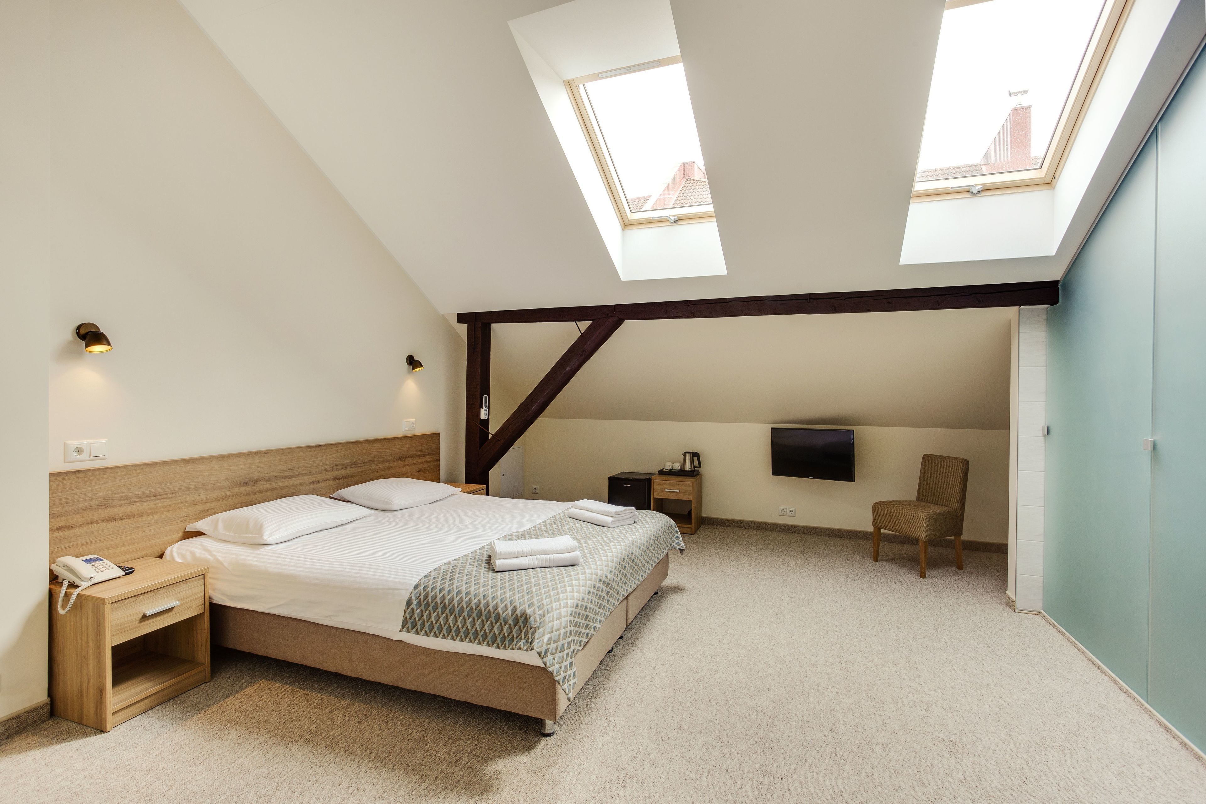 A bedroom with a skylight, a bed, a nightstand, a chair, and a TV mounted on the wall.