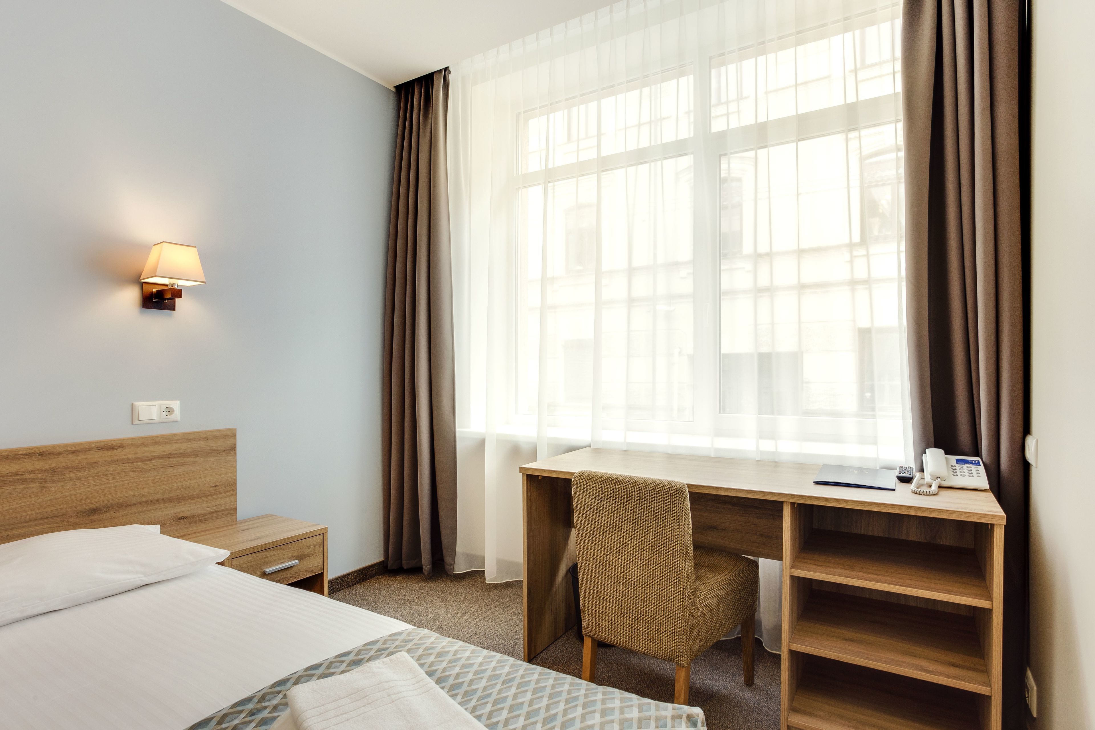 A hotel room with a bed, a desk with a chair, a lamp, a telephone, and a window with curtains.
