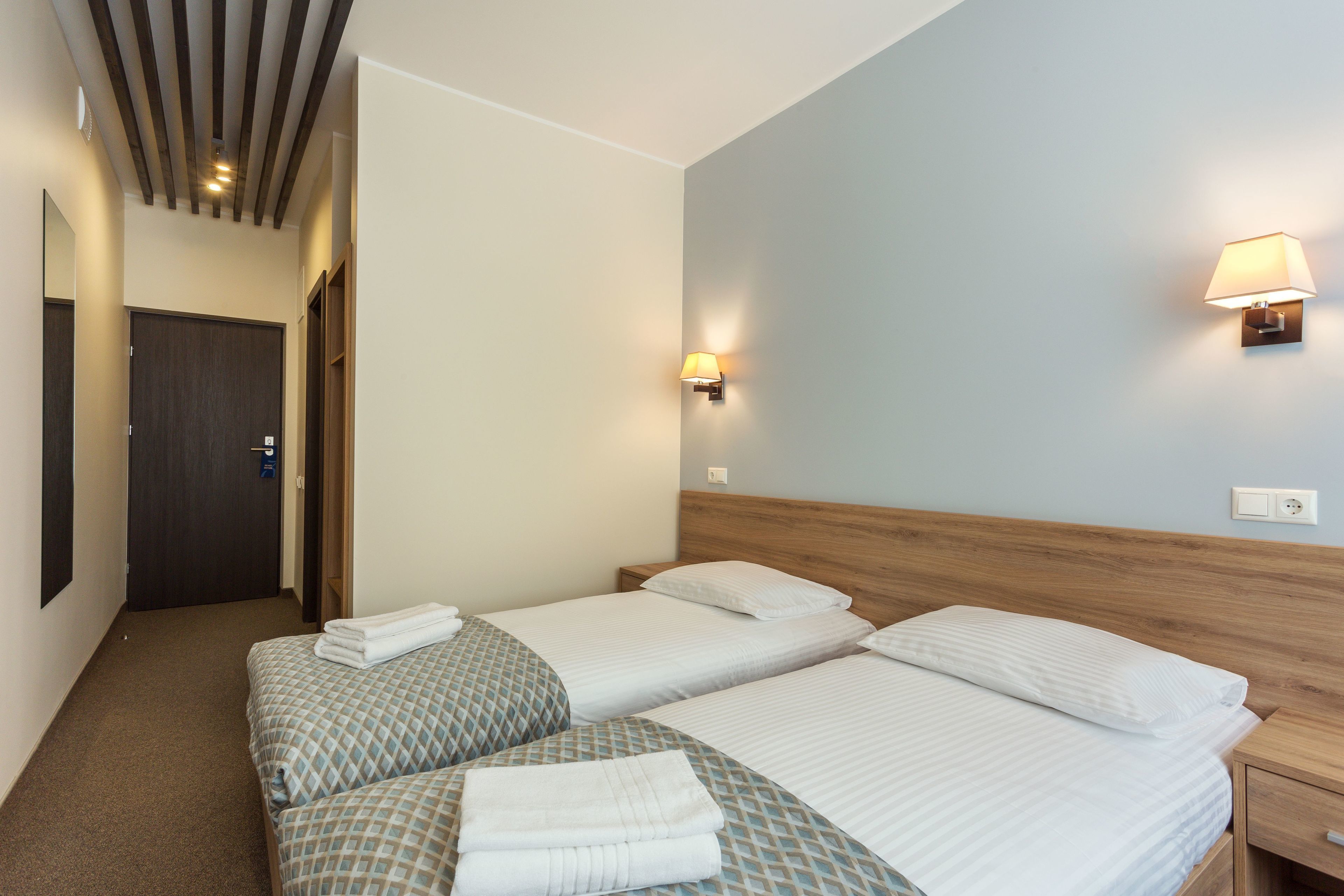 A hotel room with two beds, a wooden headboard, a wall-mounted lamp, and a door.