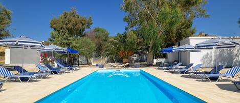 Una piscina rectangular con agua azul, rodeada de sillas de descanso y sombrillas, con árboles al fondo.