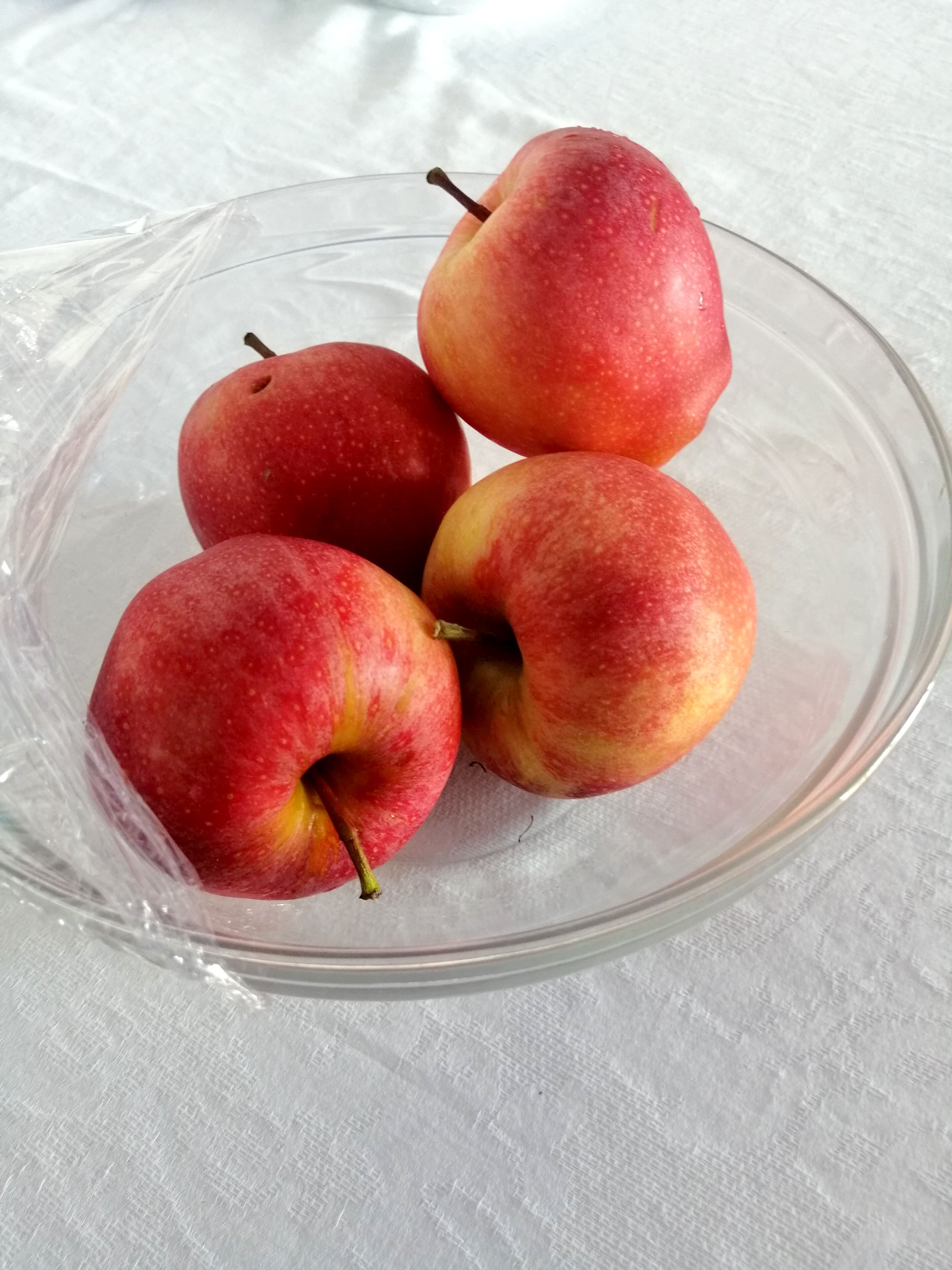 A clear glass bowl containing four red apples with yellowish hues, placed on a white textured surface.