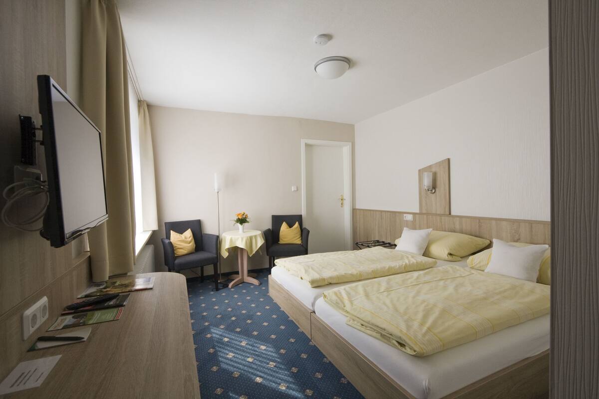 A hotel room with a bed, a desk with a chair, a small table with a vase, and a mirror on the wall.