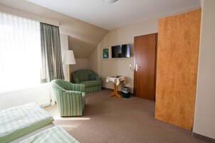 A small, well-lit room with a bed, a green-striped armchair, a small table with a lamp, a television on the wall, and a wooden door.