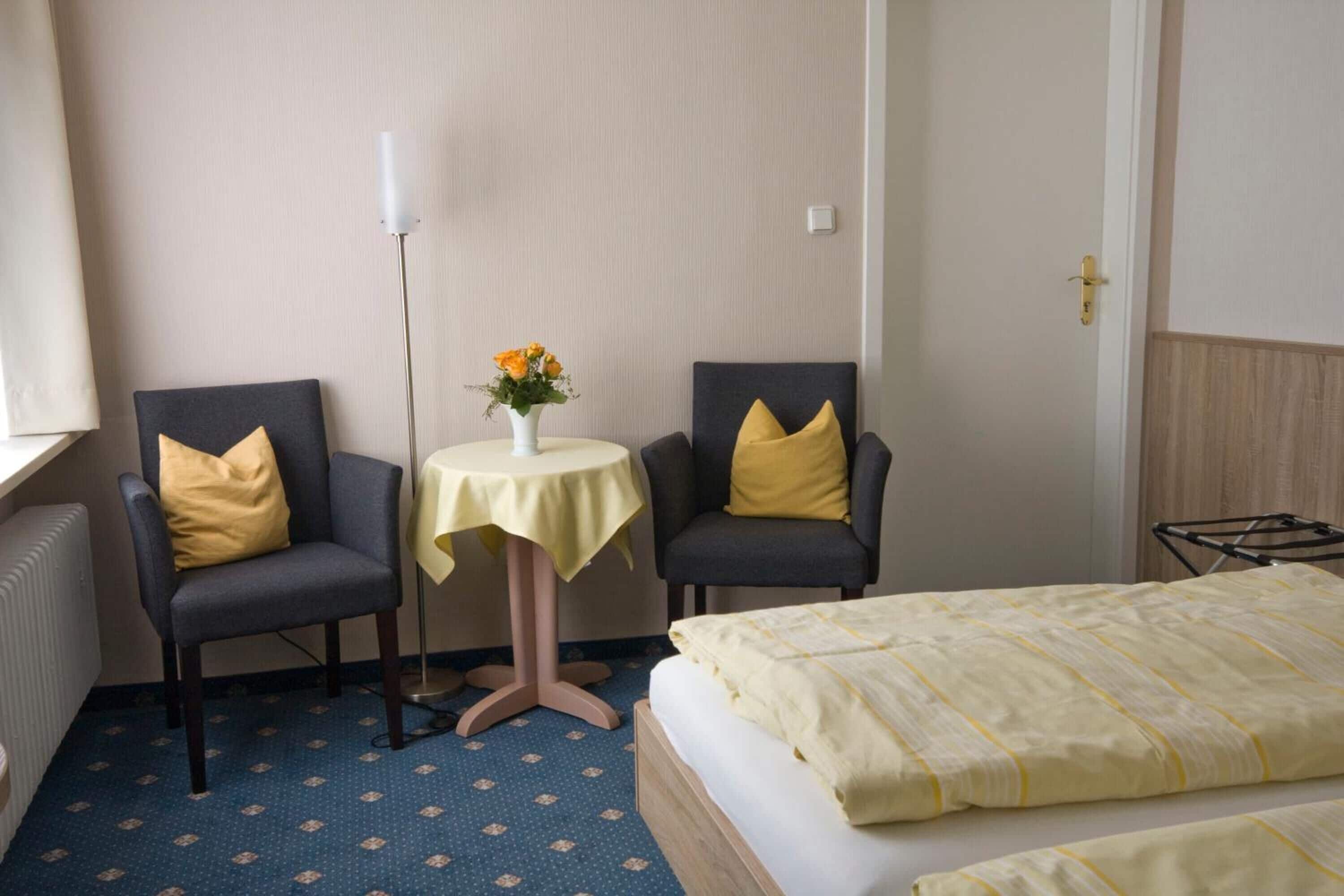 A hotel room with a bed, two armchairs, a small table with a vase of flowers, and a radiator.
