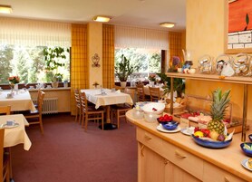 A dining area with tables set for breakfast, a fruit bowl on the counter, and a view of the outdoors through large windows.