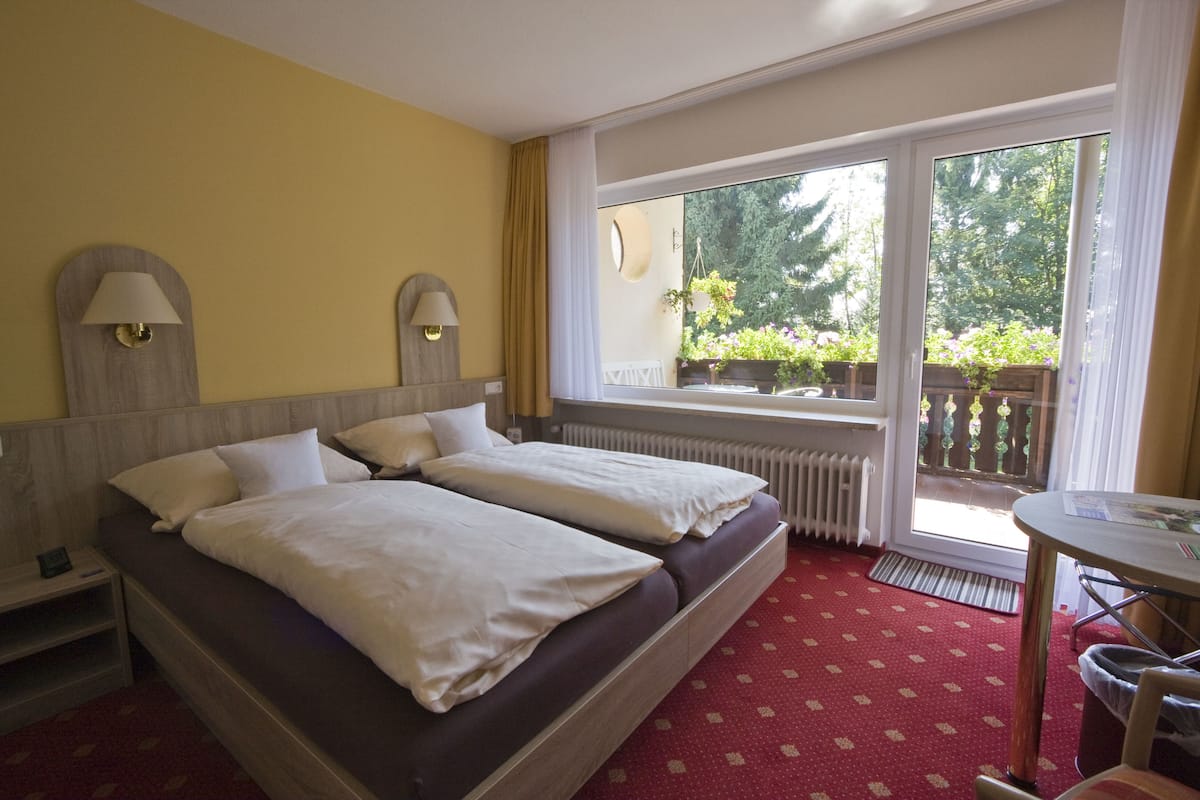 A hotel room with two beds, a small table, a window with a view of greenery, and a radiator.