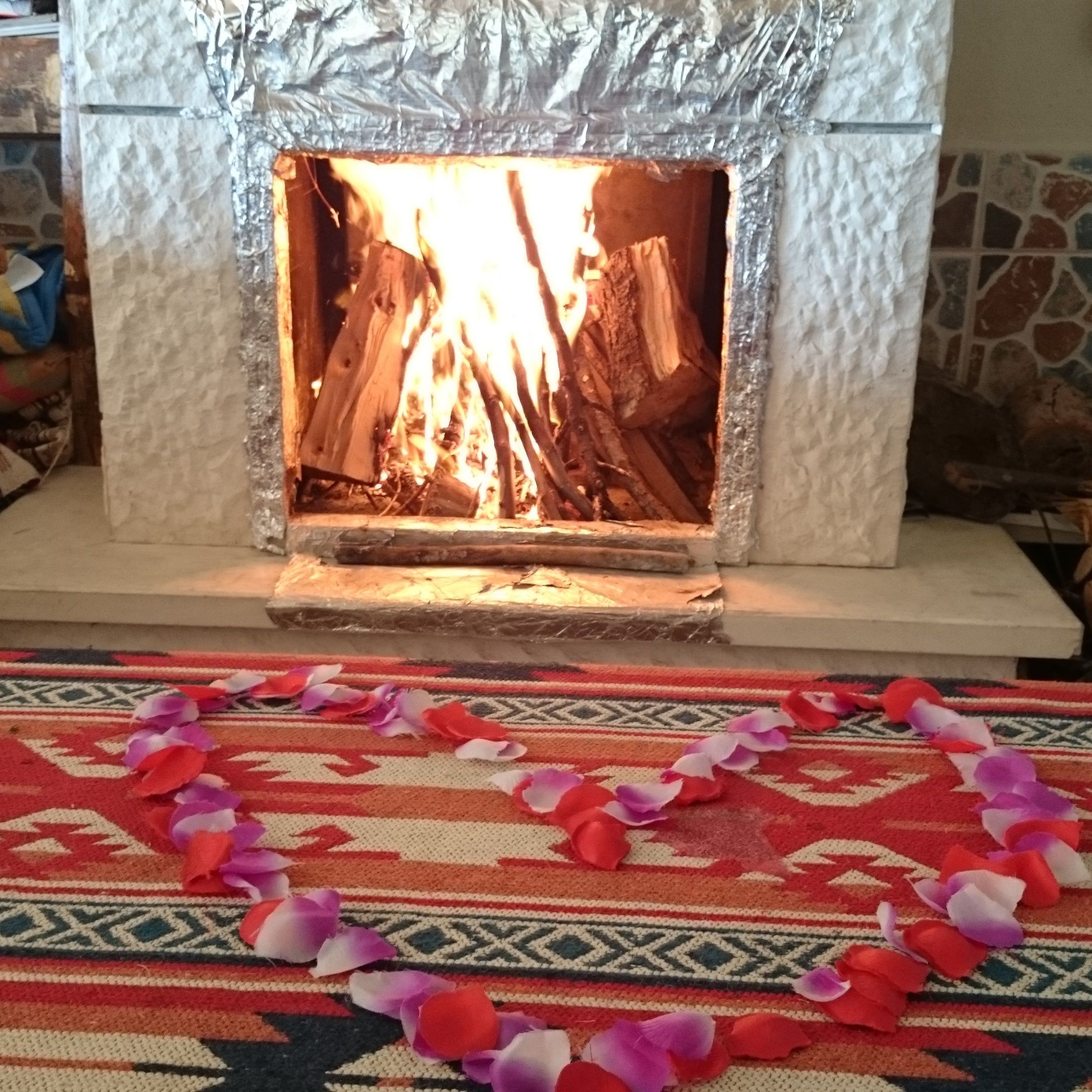 A fireplace with a fire burning inside, surrounded by a patterned rug with scattered rose petals.
