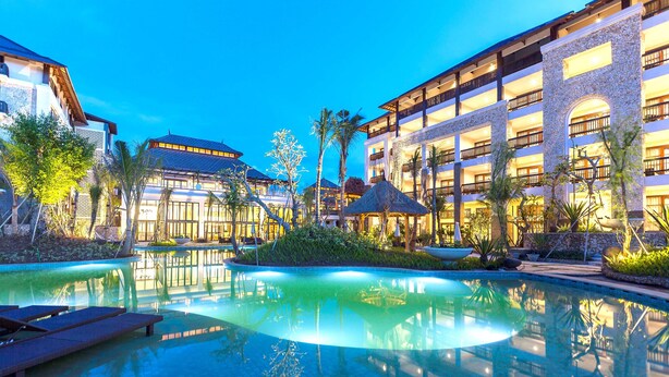 A resort pool area with lounge chairs, a water feature, and a building with multiple balconies and a stone facade. - Royal Tulip Springhill Resort Jimbaran (Jimbaran)