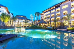 A resort pool area with lounge chairs, a water feature, and a building with multiple balconies and a stone facade. - Royal Tulip Springhill Resort Jimbaran (Jimbaran)