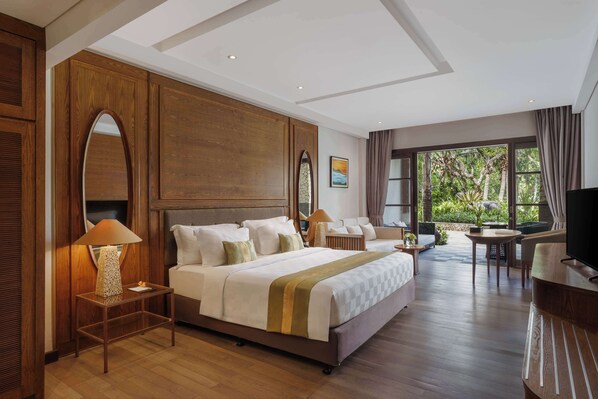 A spacious hotel room with a large bed, a sitting area with a TV, and a view of greenery through the windows. - Royal Tulip Springhill Resort Jimbaran (Jimbaran)