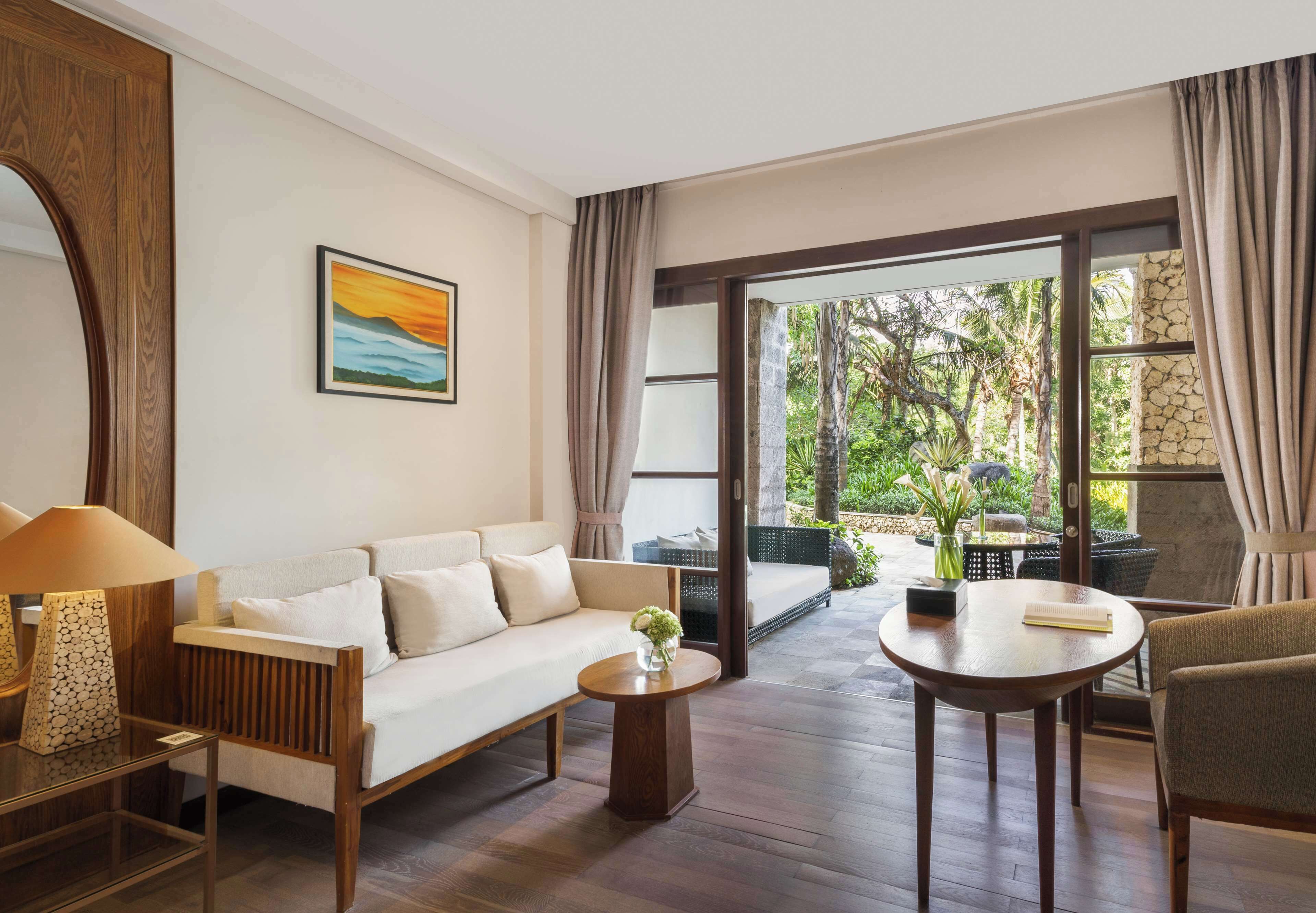 A modern living room with a sofa, coffee table, and chairs, a painting on the wall, and a view of greenery outside.