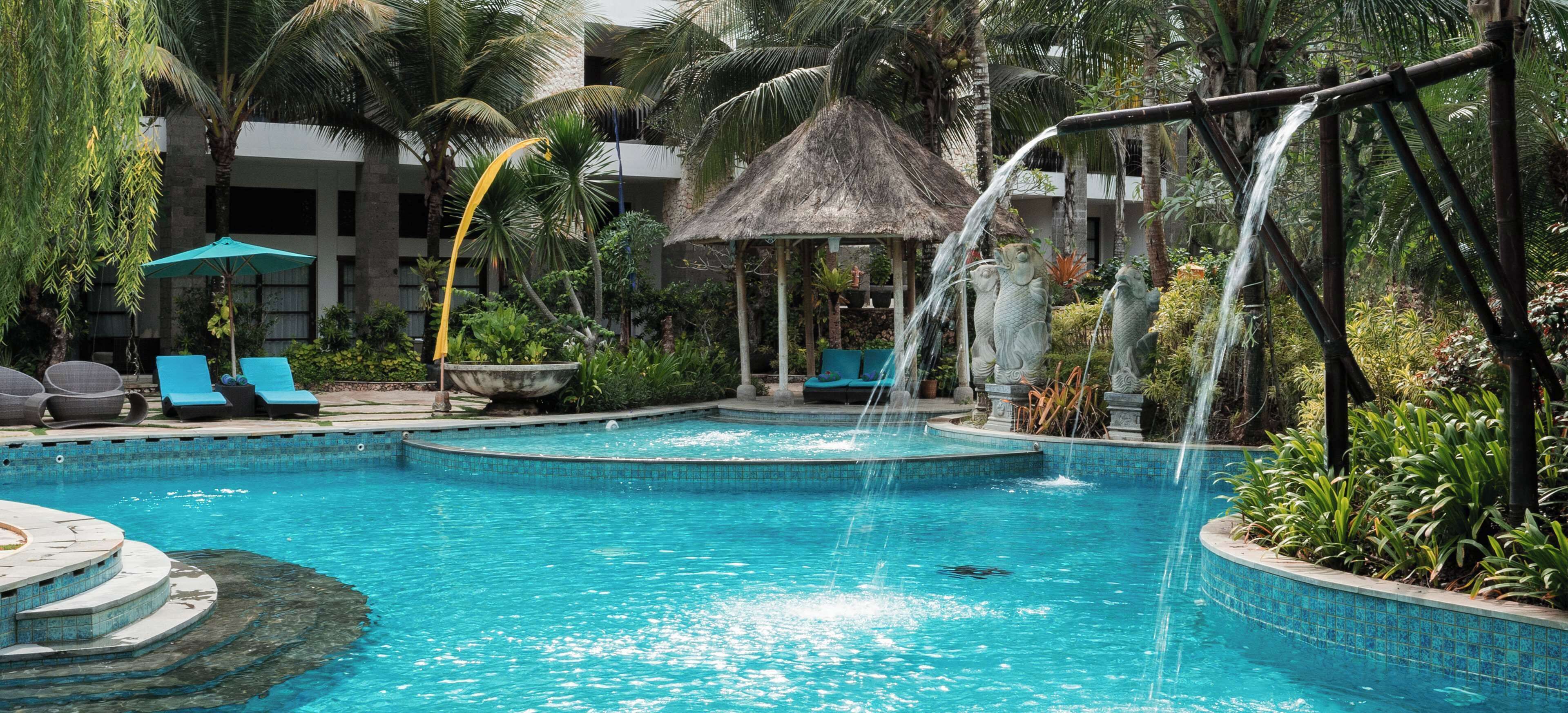 Una piscina con cascada, sillas de descanso y una estructura con techo de paja.