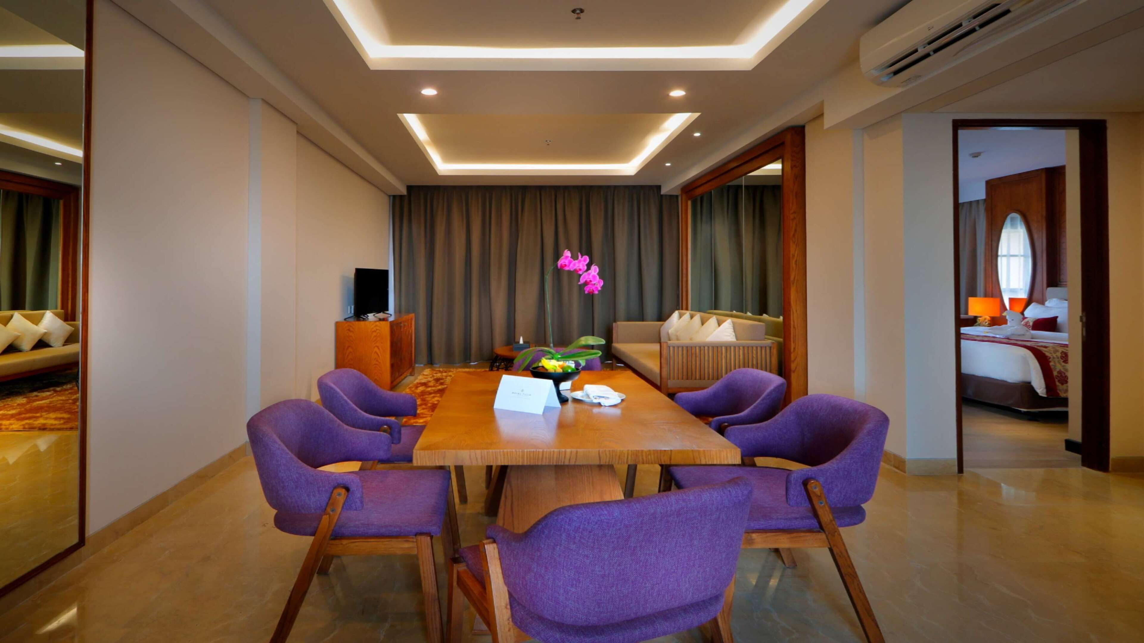 A hotel room with a large wooden dining table surrounded by purple chairs, a sofa, and a bed with a red and white bedspread.
