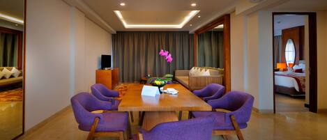 A hotel room with a large wooden dining table surrounded by purple chairs, a sofa, and a bed with a red and white bedspread.