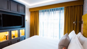 Habitación de hotel con una cama grande, un televisor de pantalla plana, un estante empotrado con adornos y un amplio ventanal con cortinas.
