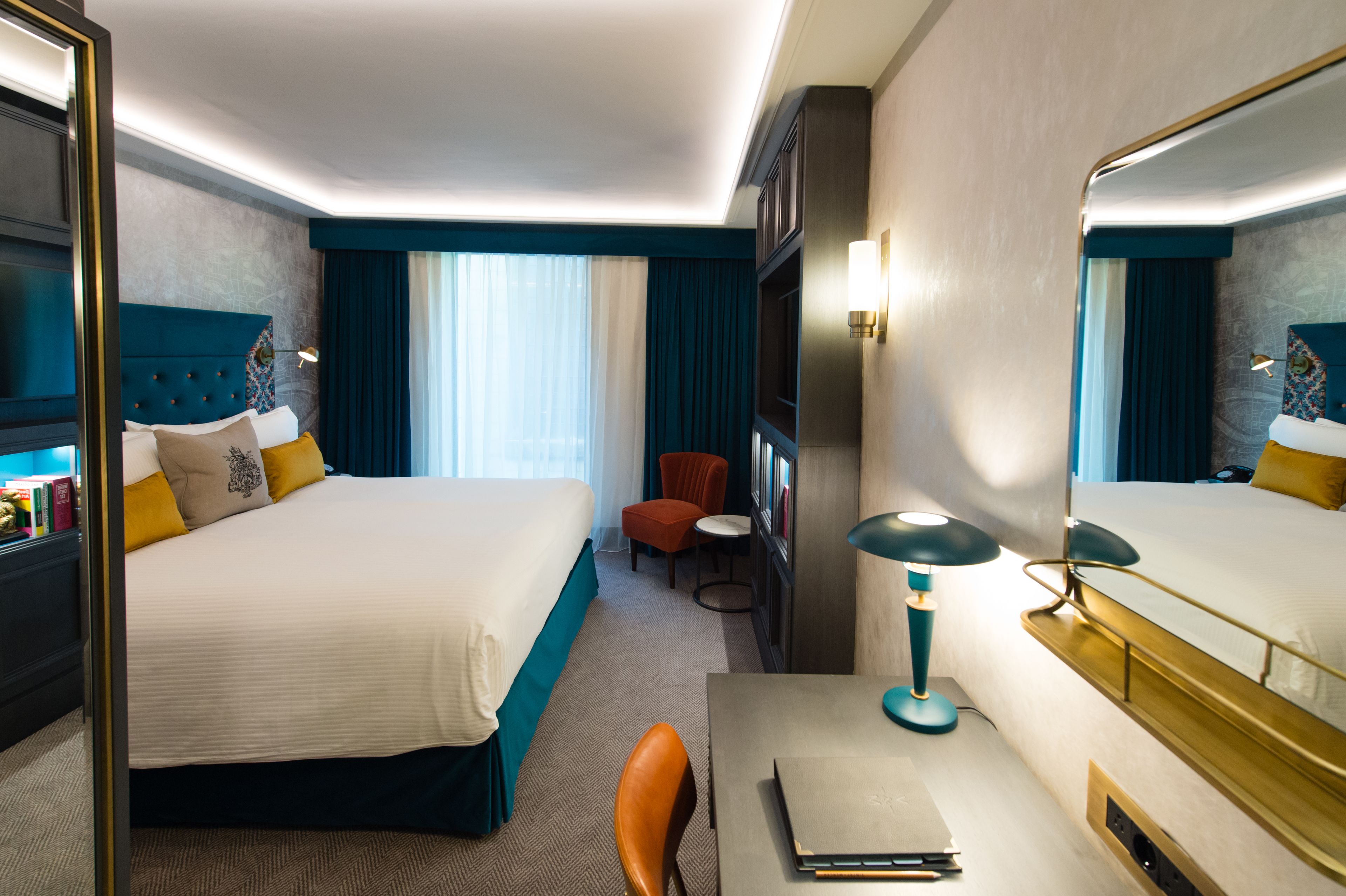Habitación de hotel con una cama grande, un escritorio con lámpara, una silla, una mesita y una estantería.