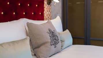 A bed with white linens and multiple pillows, including a decorative pillow with a black embroidered design. The headboard is red velvet with a tufted design and small lights.