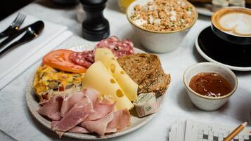 Prato de café da manhã com diversos alimentos, incluindo queijo, presunto e pão, além de uma xícara de café e uma tigela de granola.