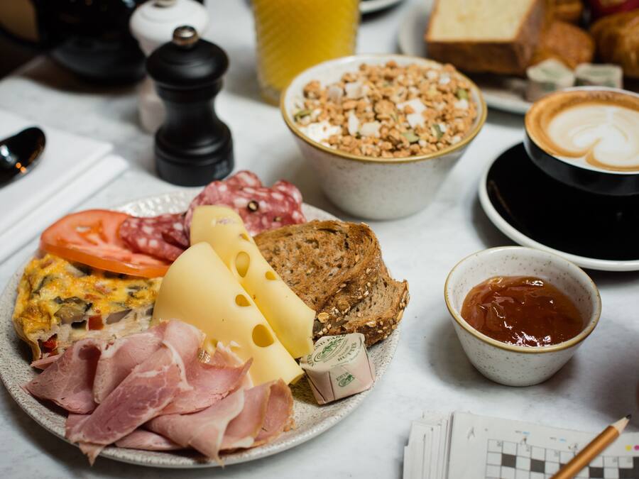 Prato de café da manhã com diversos alimentos, incluindo queijo, presunto e pão, além de uma xícara de café e uma tigela de granola.