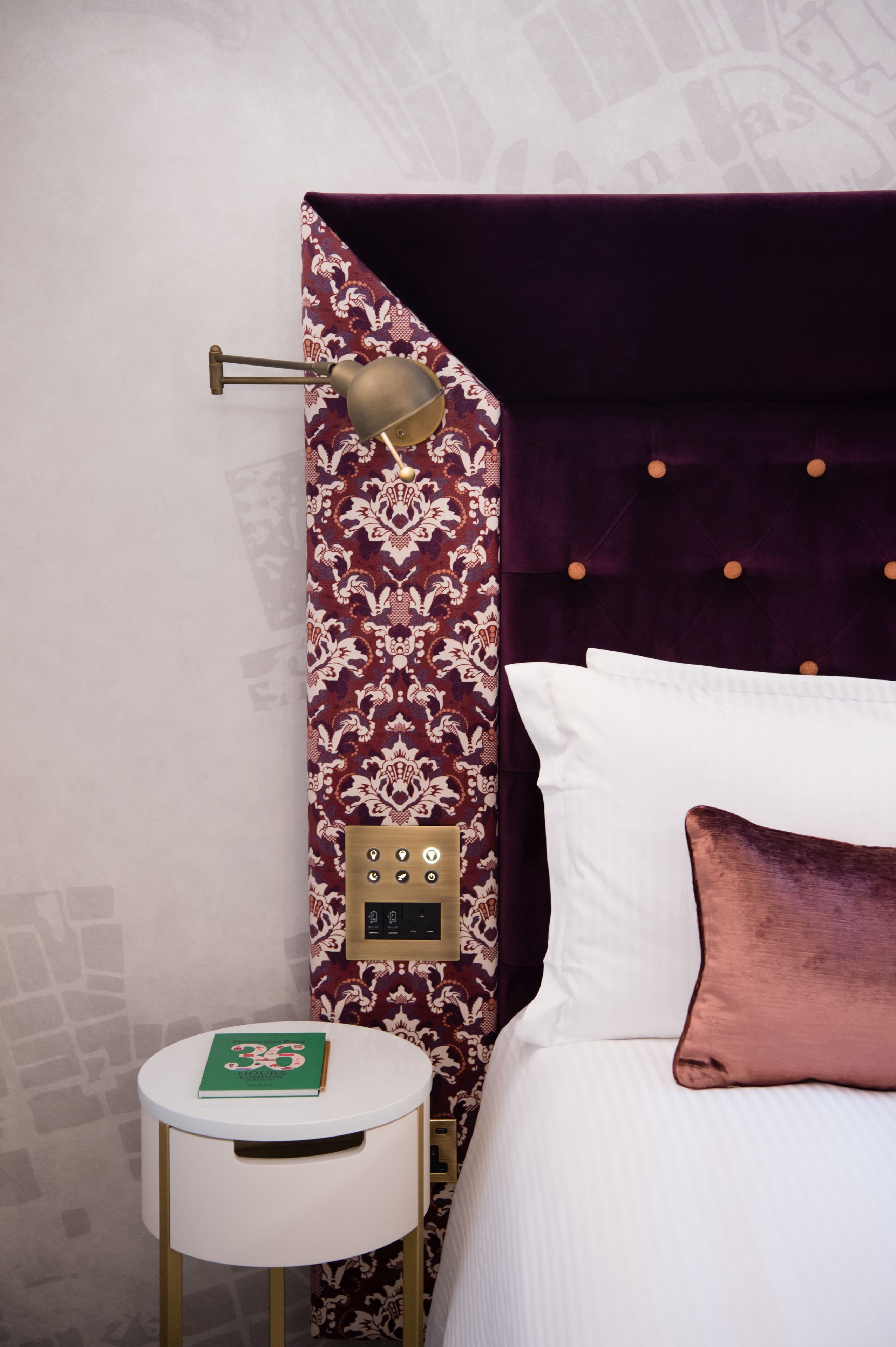 A hotel room with a patterned wallpaper, a round white table with a green book, a bed with white and pink pillows, and a modern headboard.