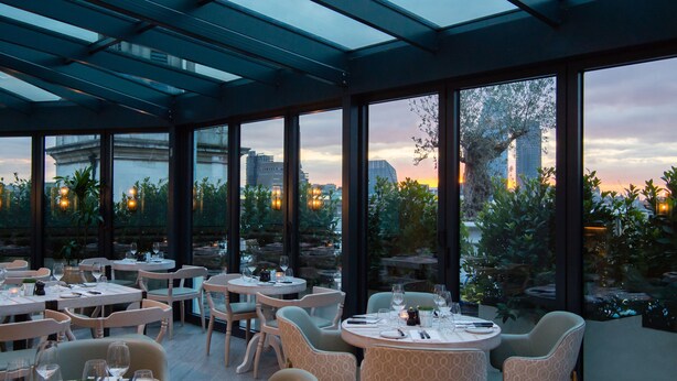 Un restaurante con mesas y sillas redondas, grandes ventanales con vistas a un paisaje urbano y al atardecer, y una estructura de techo de cristal.
