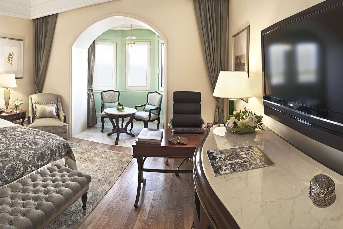 A hotel room with a large bed, a desk with a laptop, a TV, and a seating area with chairs.