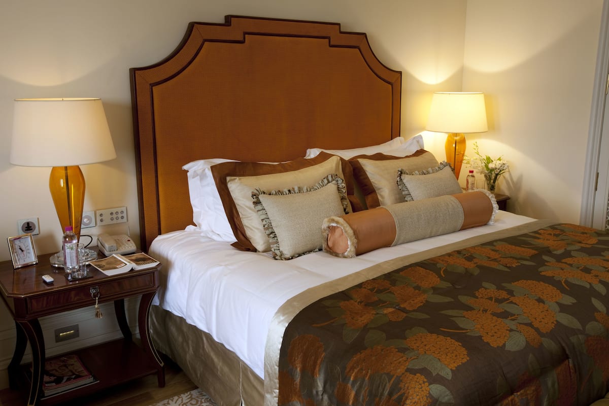 A hotel room with a large bed, two bedside tables with lamps, a nightstand, and a small table with a clock and a bottle.