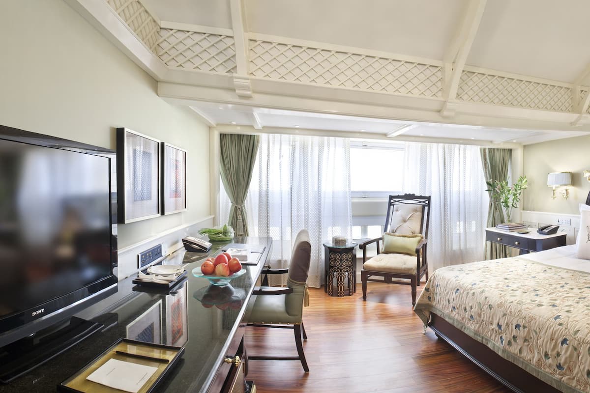 A hotel room with a large flat-screen TV, a dining area with a table and chairs, a bed with a floral bedspread, and a nightstand with a lamp.