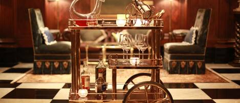 A bar cart with glasses, bottles, and a wheel in a room with checkered flooring and two lamps.