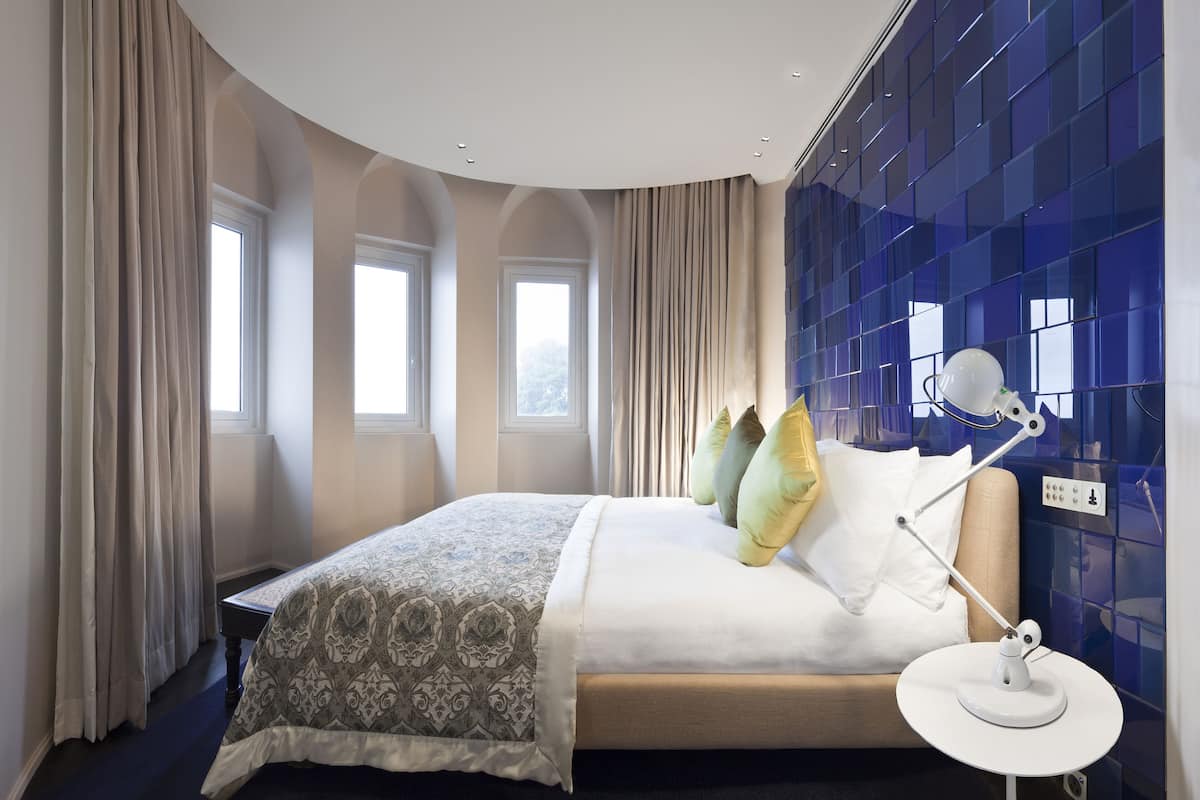 A modern hotel room with a large bed, a bedside table with a lamp, and a view of the city through the windows.