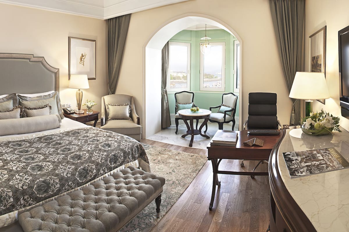 A hotel room with a large bed, a desk with a laptop, a TV, and a view through an arched doorway.