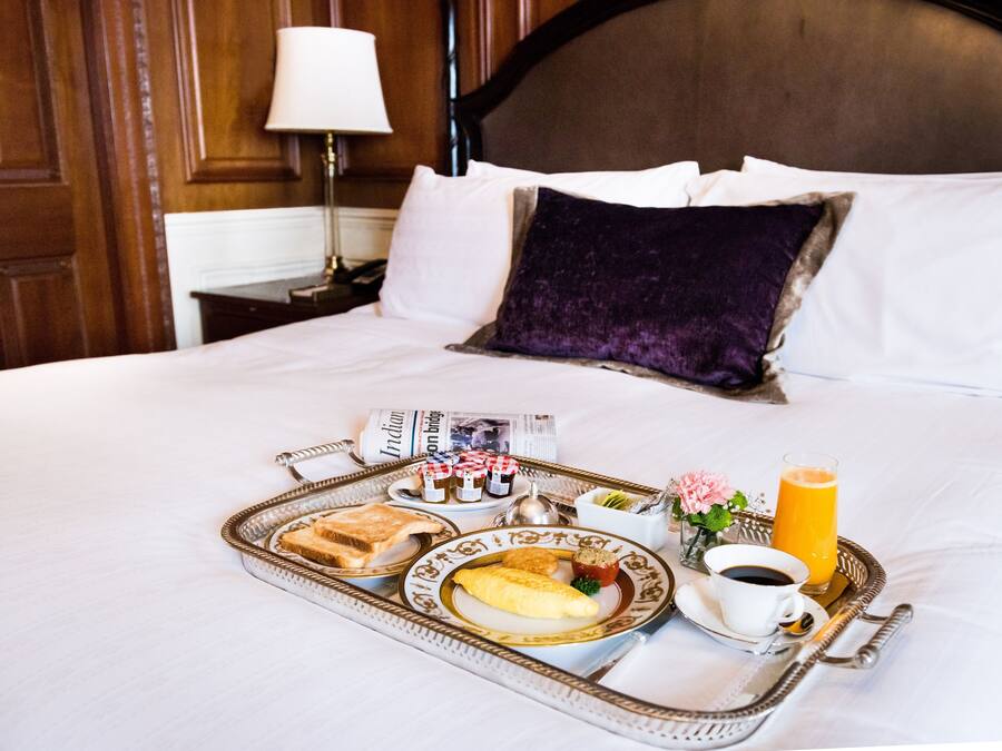 Una cama bien tendida con sábanas blancas y una almohada morada, una bandeja con alimentos para el desayuno, incluyendo tostadas, huevos y jugo, y un periódico sobre la cama.